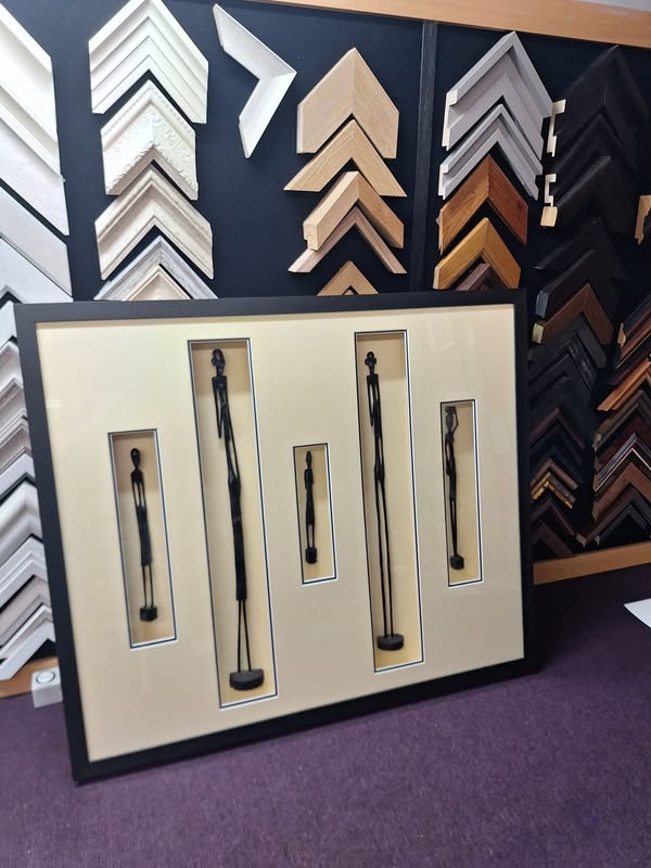 Five slender carved figurines individually mounted in a black display frame with cream mount